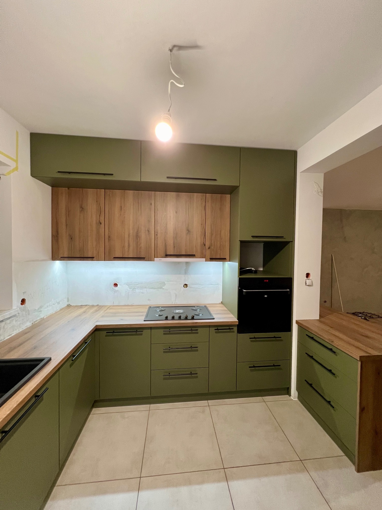 Nowoczesna kuchnia w odcieniach zieleni z drewnianymi akcentami, czarnymi uchwytami i blatem, widoczne niedokończone prace wykończeniowe na ścianie i wisząca żarówka bez oprawy.