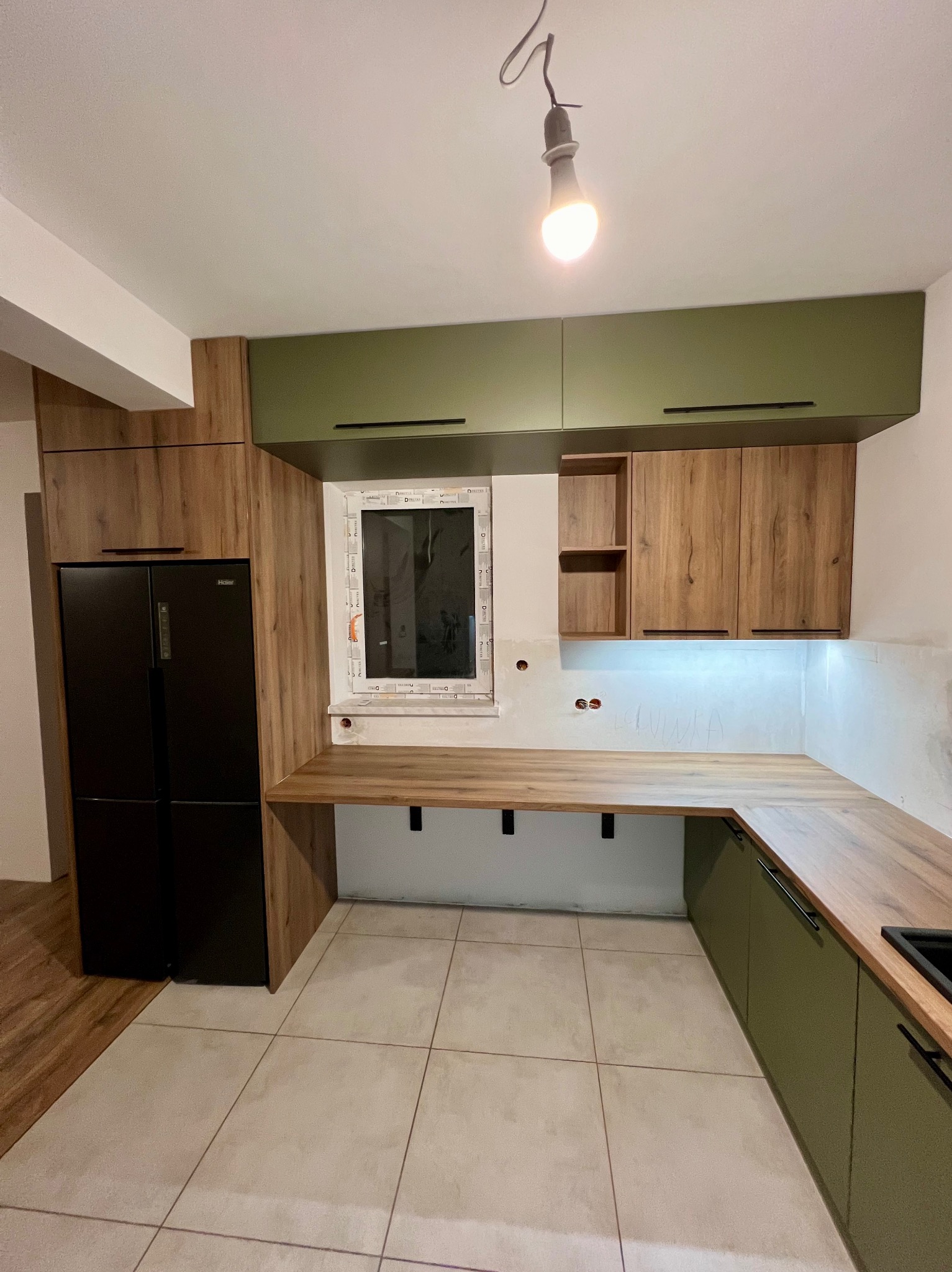 Wnętrze kuchni w trakcie montażu: zielone i drewnopodobne szafki górne i dolne z czarnymi uchwytami, drewniany blat roboczy pod oknem, czarna lodówka side-by-side, surowa ściana z widocznymi...
