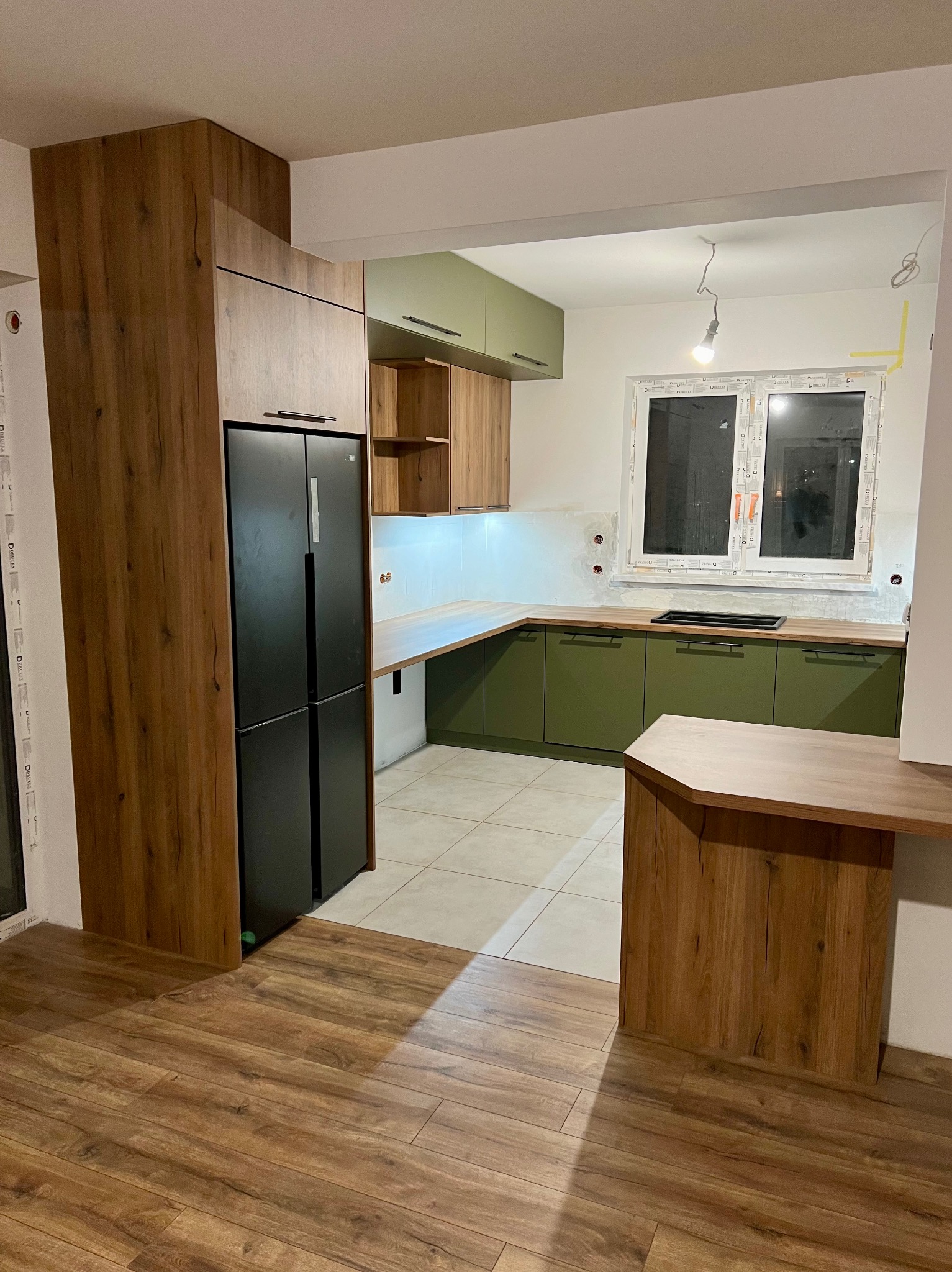 Nowoczesna kuchnia z zielonymi szafkami dolnymi, drewnianymi szafkami górnymi i wysoką zabudową z lodówką w wykończeniu drewnopodobnym, widok na fragment blatu i okno.