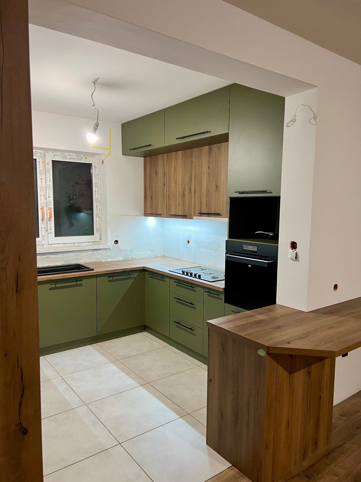 Montaż kuchni: zielone i drewniane szafki, czarny piekarnik, widoczne niedokończone prace wykończeniowe przy oknie i ścianie.