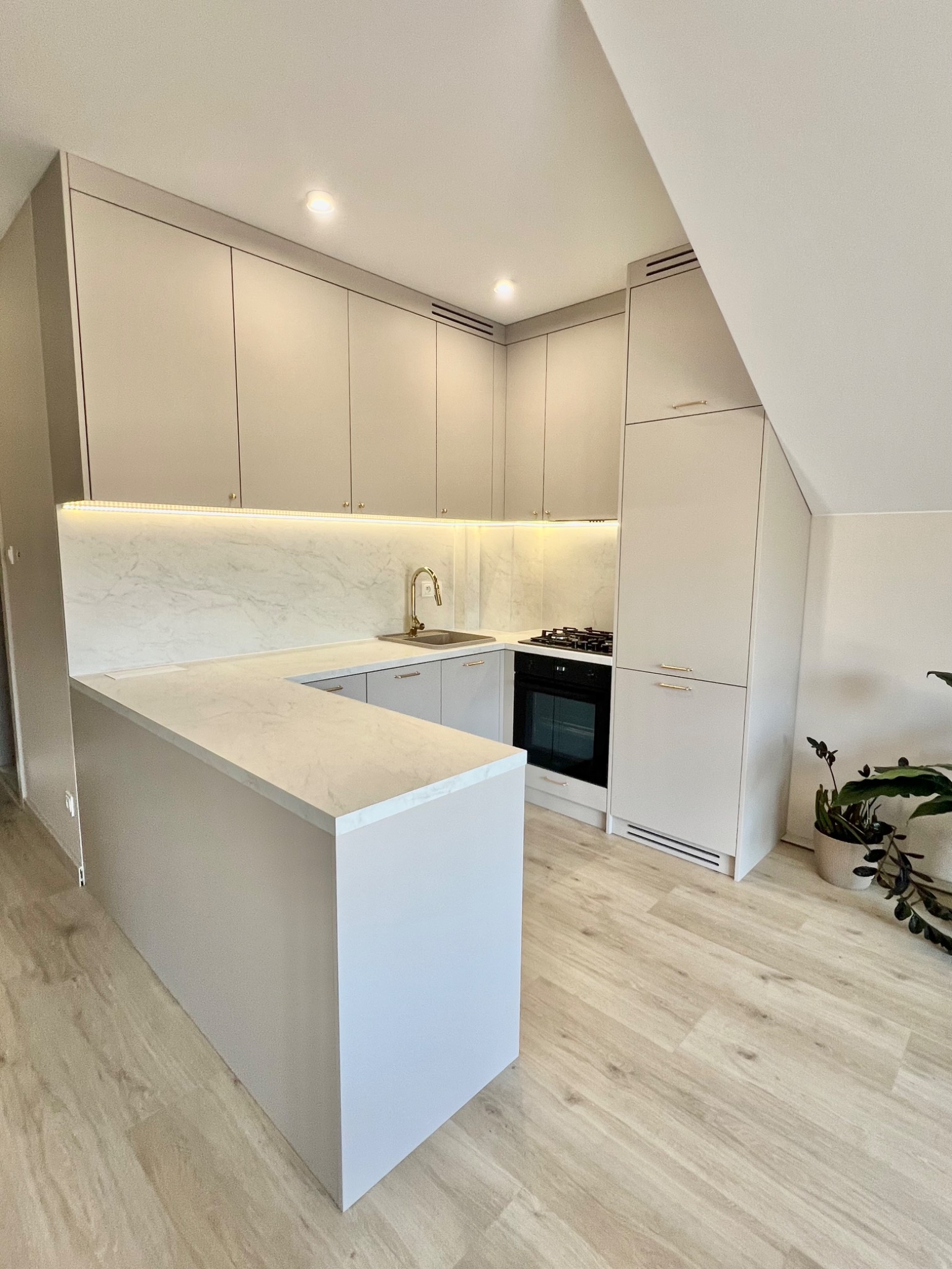 Jasna kuchnia z zabudową pod skosem, marmurowy blat i złota bateria, oświetlenie LED pod szafkami, drewnopodobna podłoga.