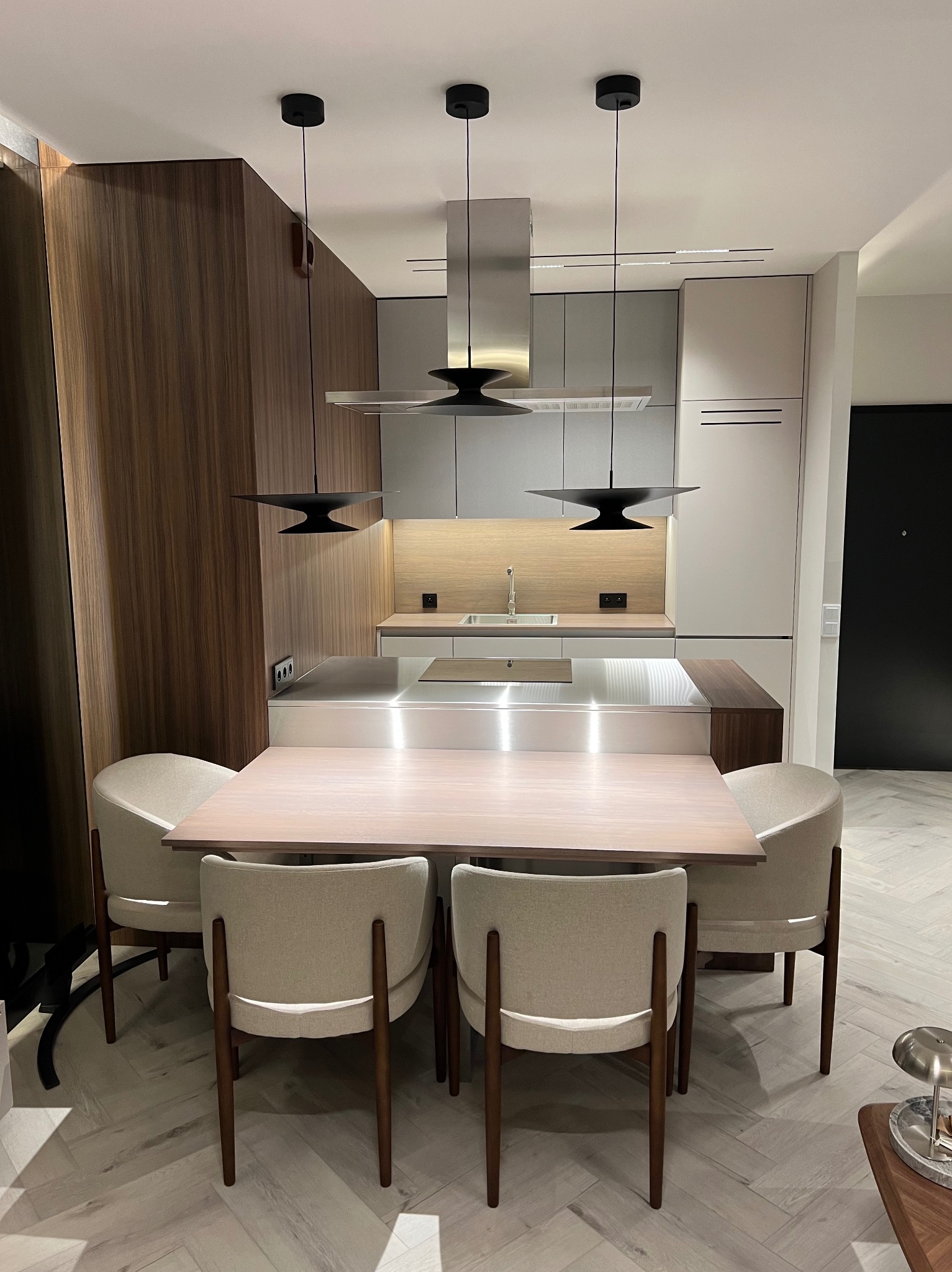 Nowoczesna kuchnia z drewnianymi frontami szafek, wyspą ze stali nierdzewnej i stołem jadalnym z krzesłami w kolorze beżowym, oświetlona trzema wiszącymi lampami w stylu industrialnym.