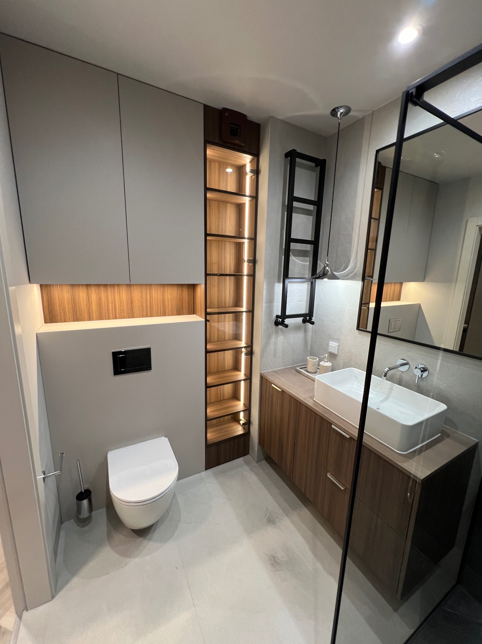 Nowoczesna łazienka z wiszącą toaletą, szafkami w odcieniach szarości i drewna, podświetlaną półką nad toaletą i otwartym regałem z oświetleniem LED, umywalka nablatowa.
