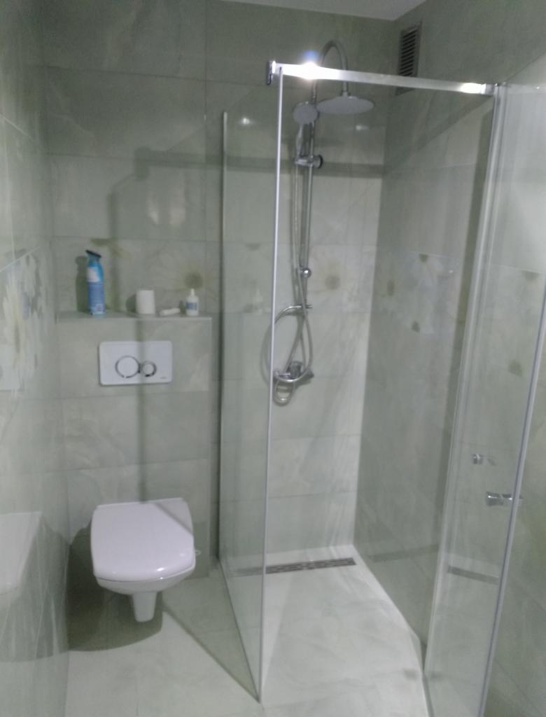 Nowoczesna kabina prysznicowa walk-in z odpływem liniowym, zamontowana w wykończonej łazience z jasnymi płytkami imitującymi marmur i podwieszaną toaletą.