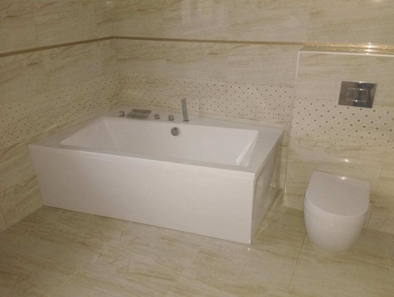 Nowoczesna, biała wanna narożna z hydromasażem i armaturą, obok toaleta, w łazience wykończonej płytkami imitującymi marmur z dekoracyjnym pasem.