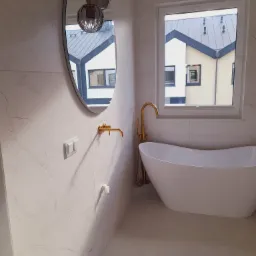 Nowoczesna łazienka z wolnostojącą wanną, złotą armaturą i owalnym lustrem, odbijającym budynek na zewnątrz.