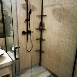 Turczyn Daniel - usługi budowlane - Nowoczesna kabina prysznicowa z czarną armaturą, beżowe płytki ścienne z delikatnym wzorem, drewnopodobna podłoga, narożna półka na kosmetyki, odbicie fotografa w szkle.
