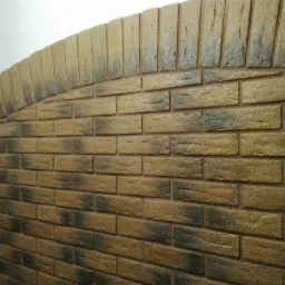 Dekoracyjna ściana z cegieł klinkierowych w odcieniach brązu i beżu, ułożona w łukowaty kształt, z widoczną fakturą i cieniowaniem cegieł.