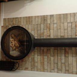 Turczyn Daniel - usługi budowlane - Nowoczesny kominek z okrągłym paleniskiem i ułożonymi w pionie płytkami imitującymi cegłę na ścianie, z widocznym drewnem wewnątrz paleniska i czarnym grillem obok.