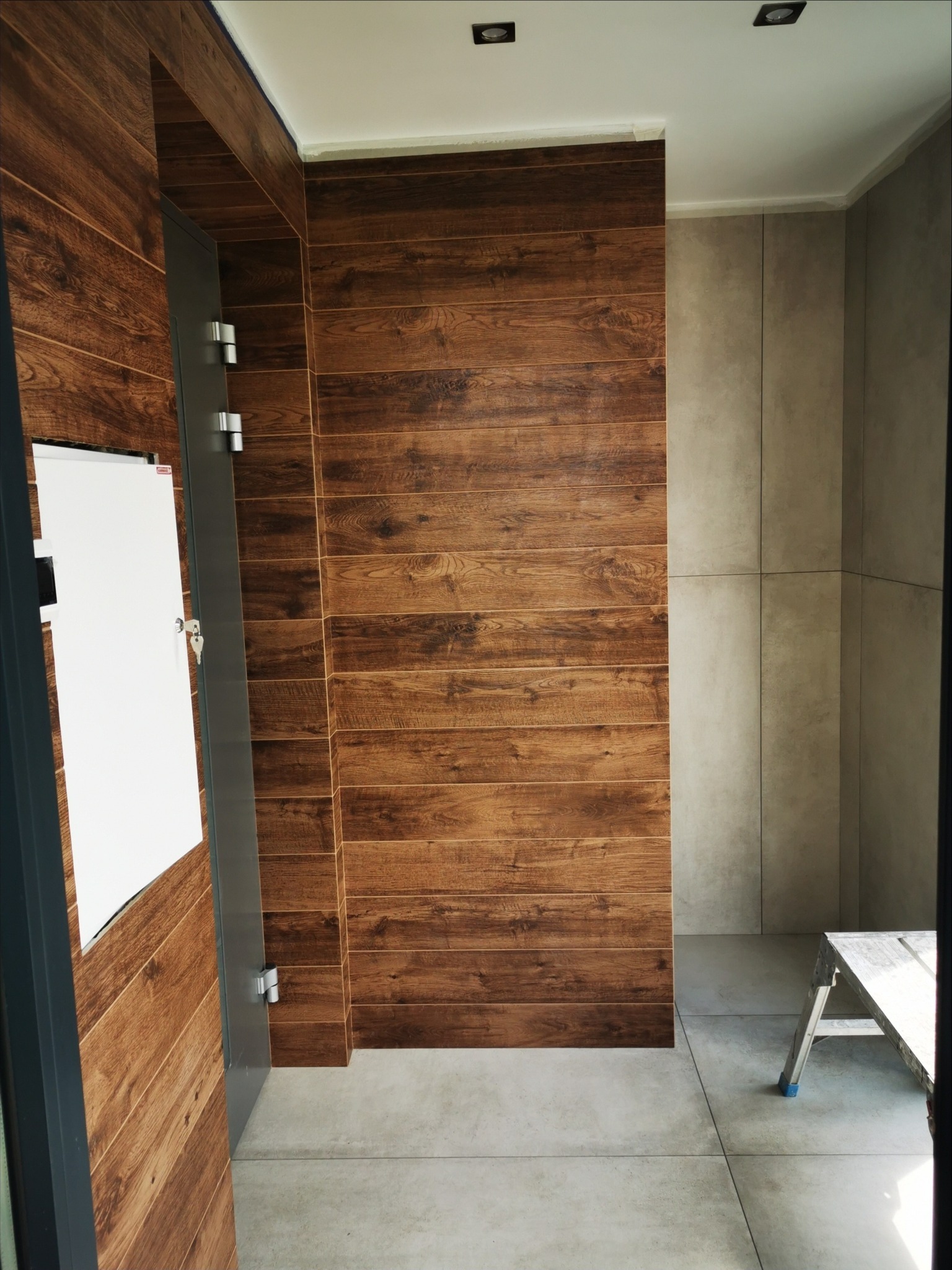 Wykończona łazienka z płytkami imitującymi drewno na ścianach i szarymi płytkami na podłodze, widoczna drabina malarska w rogu pomieszczenia i otwarte drzwi z metalowymi zawiasami.