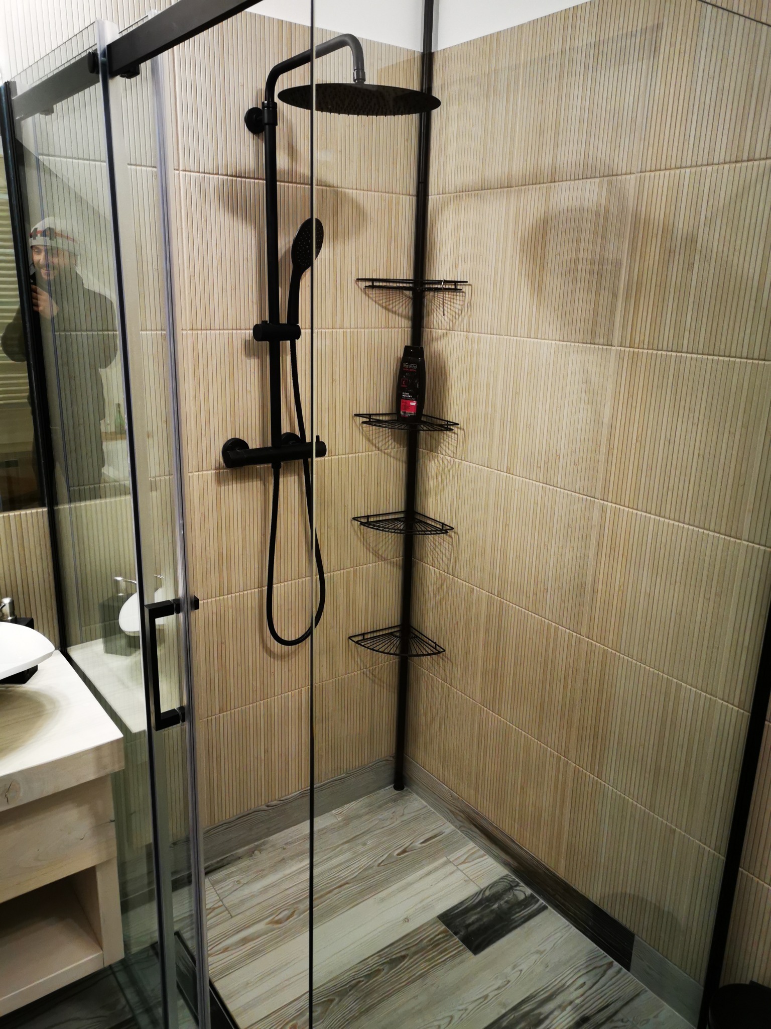 Nowoczesna kabina prysznicowa z czarną armaturą, beżowe płytki ścienne z delikatnym wzorem, drewnopodobna podłoga, narożna półka na kosmetyki, odbicie fotografa w szkle.