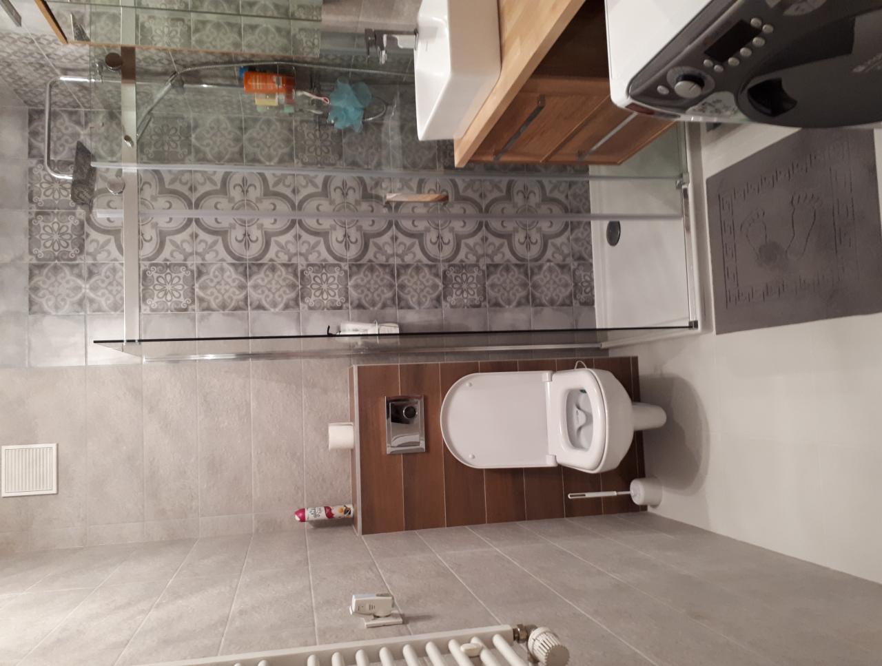 Nowoczesna łazienka z szarą płytką dekoracyjną, kabiną prysznicową ze szklaną ścianą, podwieszaną toaletą i drewnianą szafką pod umywalką, widoczny grzejnik drabinkowy.