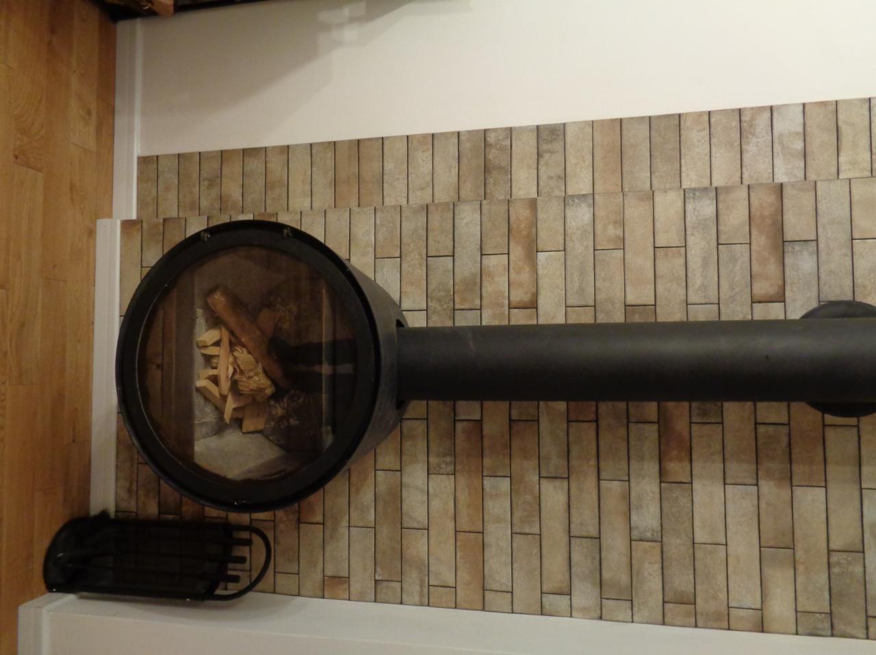 Nowoczesny kominek z okrągłym paleniskiem i ułożonymi w pionie płytkami imitującymi cegłę na ścianie, z widocznym drewnem wewnątrz paleniska i czarnym grillem obok.