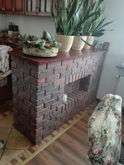 Wykończona ściana z cegły klinkierowej tworząca barek kuchenny z wnęką, na blacie dekoracja świąteczna i doniczki z roślinami, widok z boku na tle kuchni.