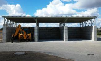 Wiata z prefabrykatów betonowych z żółtą koparką wewnątrz, widoczne 3 segmenty zadaszone blachą trapezową, kostka brukowa na placu.