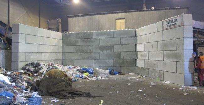 Ściana boksów z szarych betonowych bloków, z napisem 'BETBLOK SYSTEM', otoczona stertą śmieci w hali.