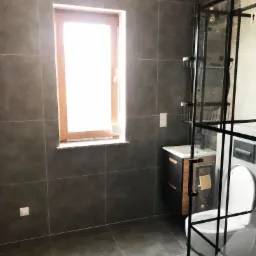 Nowoczesna łazienka z szarymi płytkami, drewnianym oknem i kabiną prysznicową z czarnymi ramami, widoczna umywalka z szafką w stylu loft.