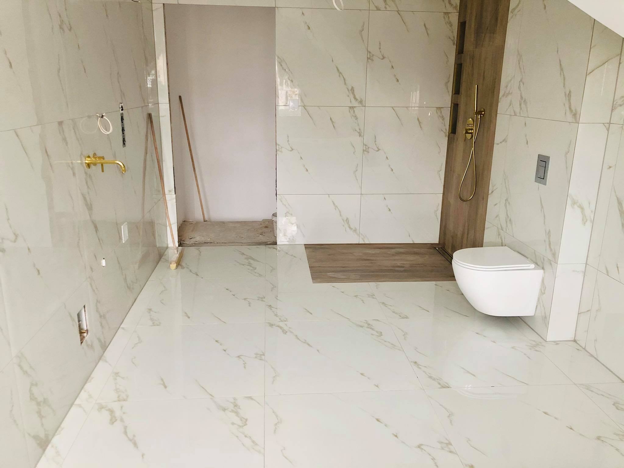 Łazienka w trakcie wykańczania: białe płytki imitujące marmur na ścianach i podłodze, złota armatura, zamontowana toaleta wisząca, widoczne przygotowania do dalszych prac.
