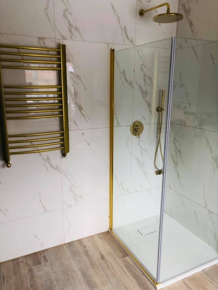 Wykończona łazienka z kabiną prysznicową z elementami w kolorze złotym, grzejnik drabinkowy w kolorze złotym, płytki imitujące marmur na ścianach i drewnopodobne płytki na podłodze.