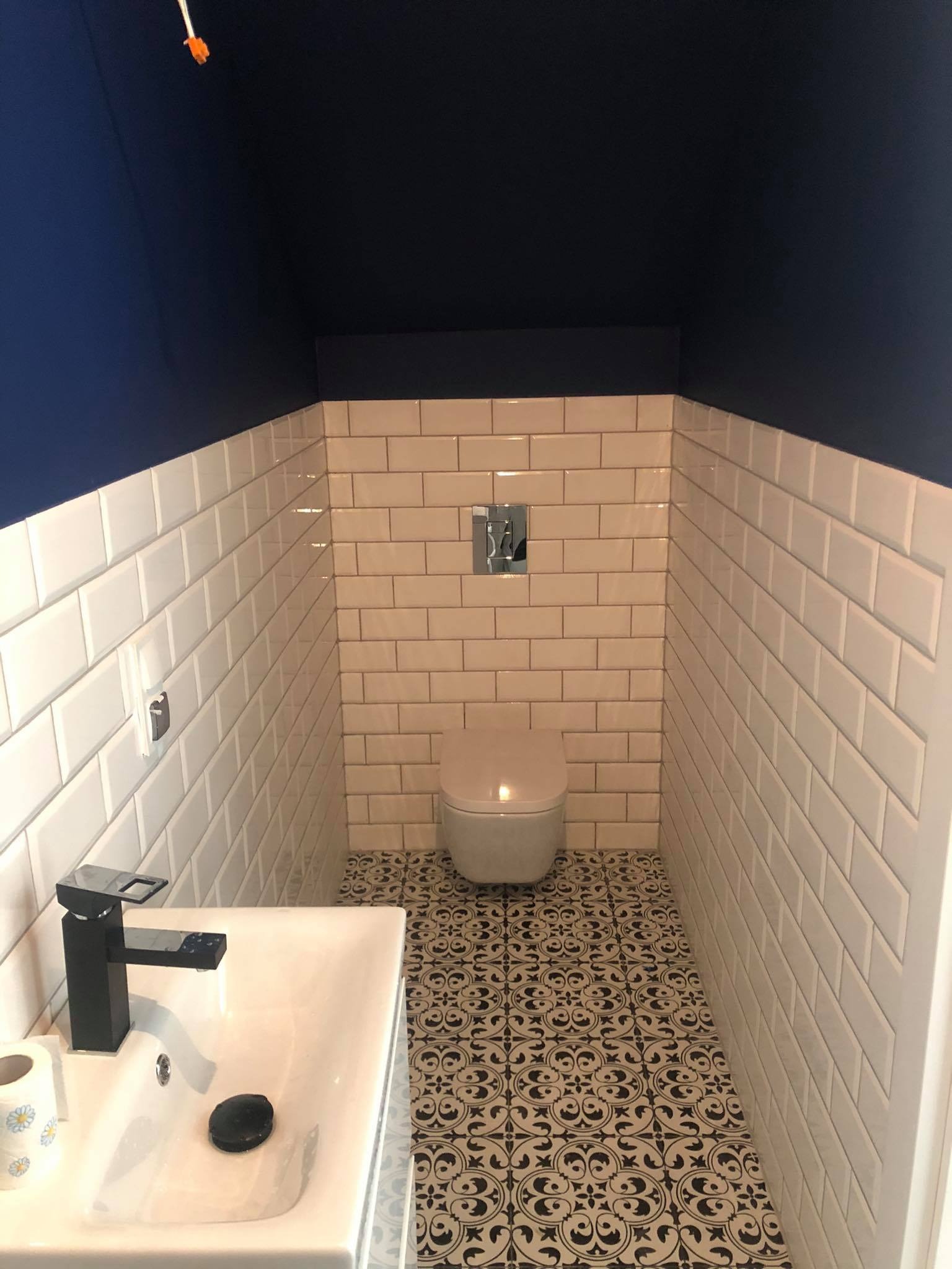 Nowoczesna toaleta z białymi, prostokątnymi płytkami, granatowym sufitem, wzorzystą podłogą i czarną armaturą przy białej umywalce