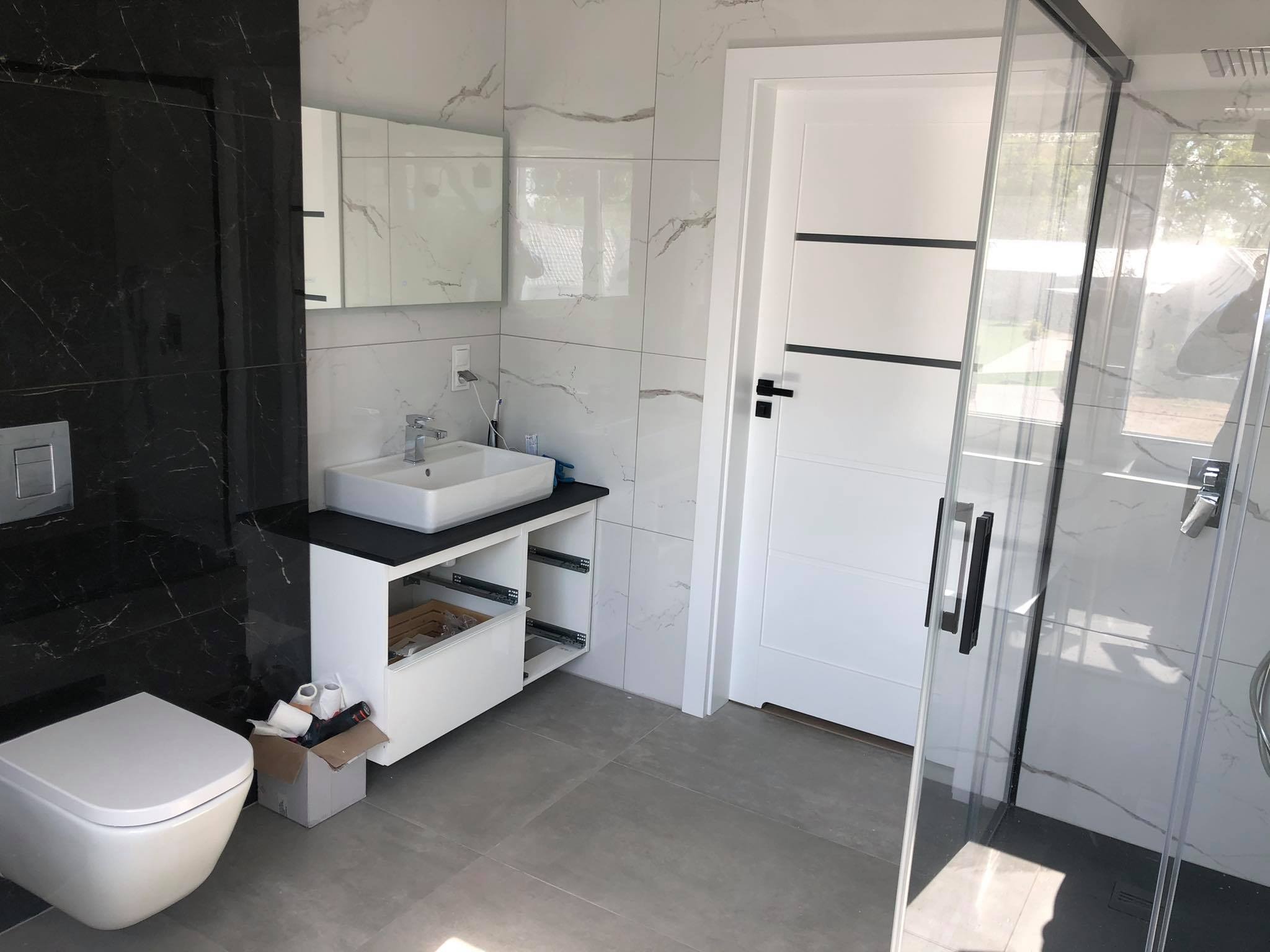 Nowoczesna łazienka z czarno-białymi płytkami imitującymi marmur, białą armaturą i kabiną prysznicową typu walk-in. Częściowo widoczny bałagan w postaci kartonu i papieru toaletowego pod umywalką.