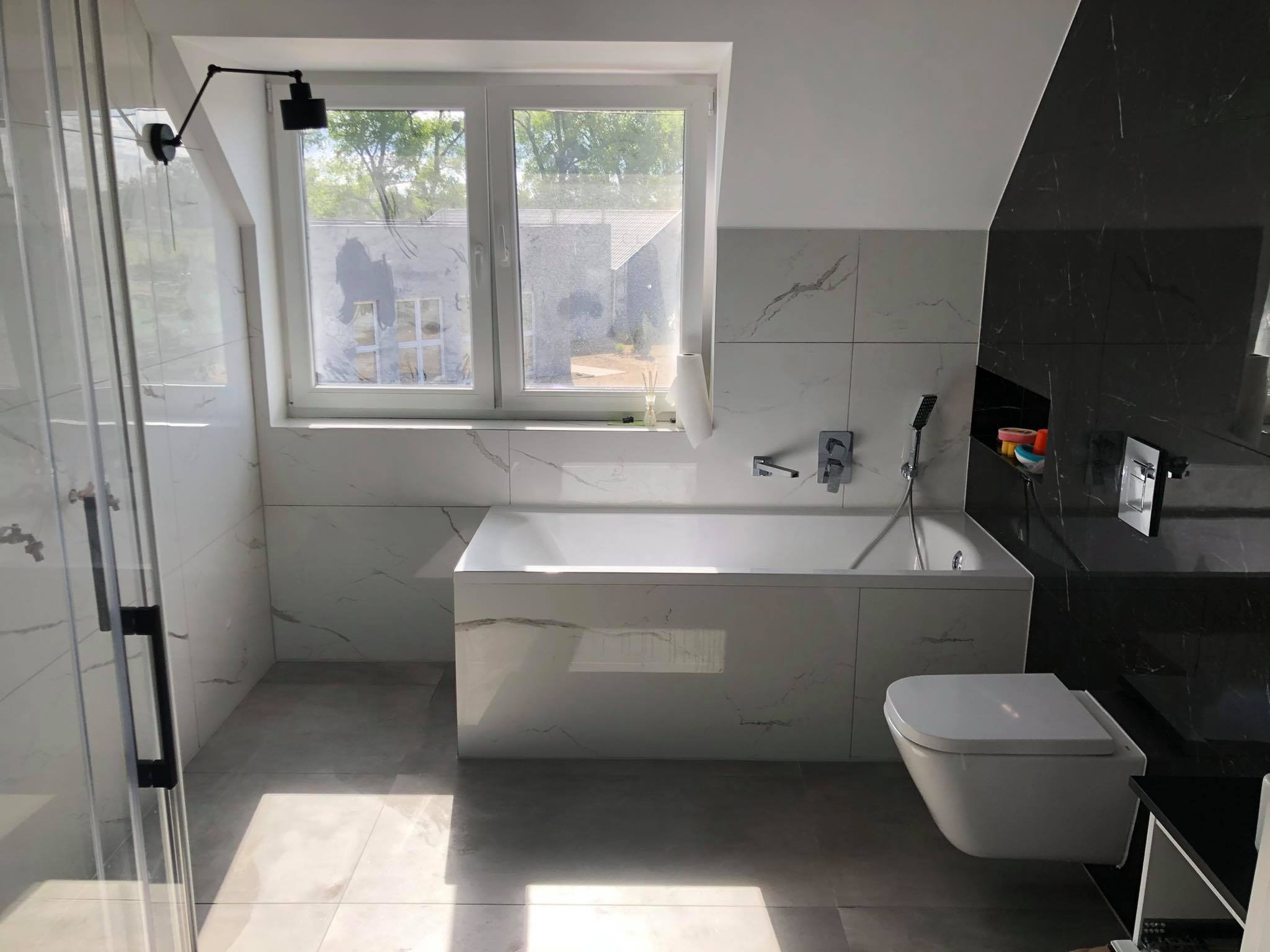 Wykończona łazienka w bieli i czerni z wanną obudowaną płytkami imitującymi marmur, wiszącą toaletą i kabiną prysznicową ze szklanymi drzwiami na tle okna z widokiem na zieleń.