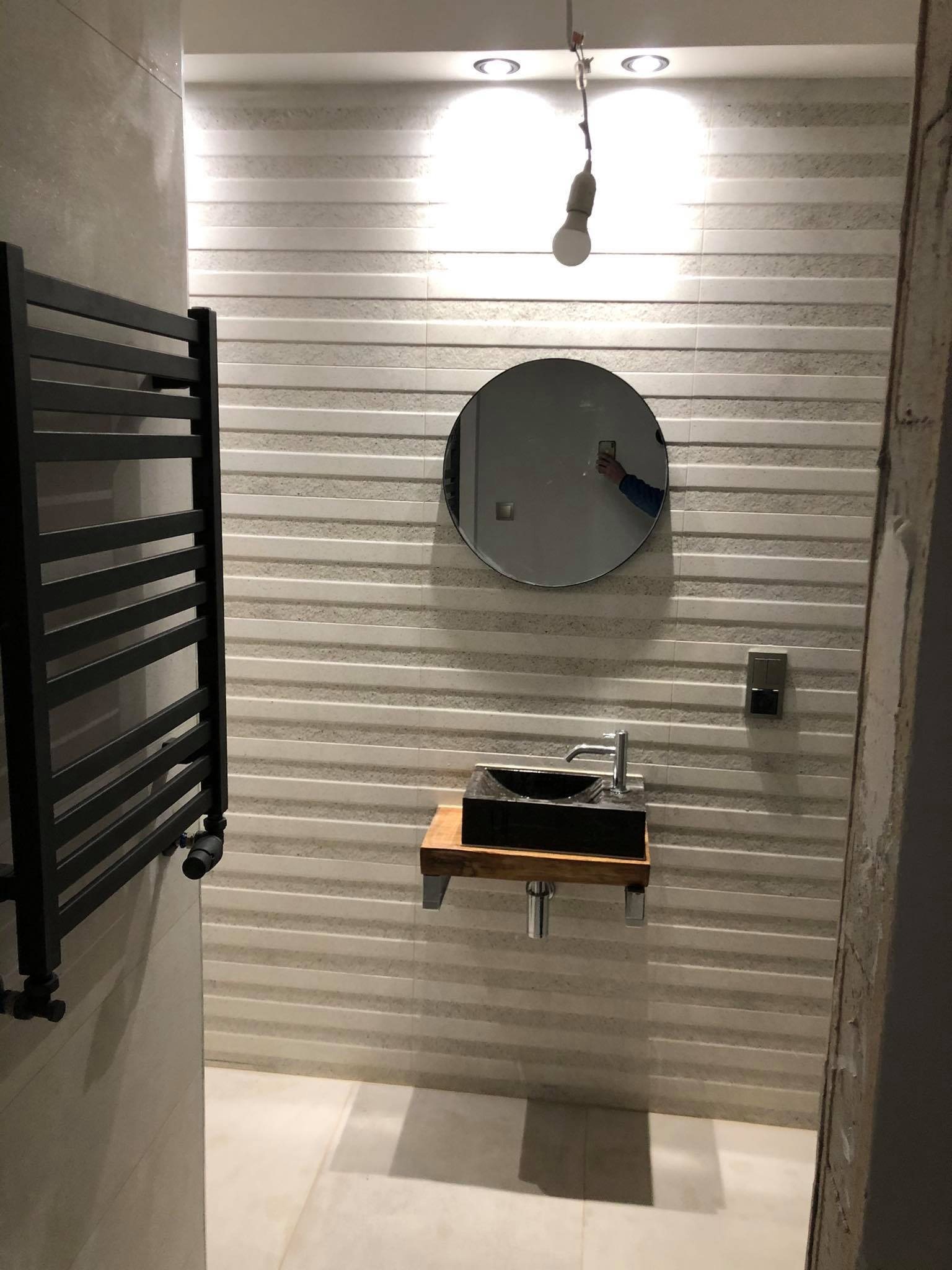 Łazienka w trakcie wykańczania: widoczny grzejnik drabinkowy, okrągłe lustro z odbiciem osoby robiącej zdjęcie, umywalka na drewnianym blacie, płytki w poziome pasy i wisząca żarówka.