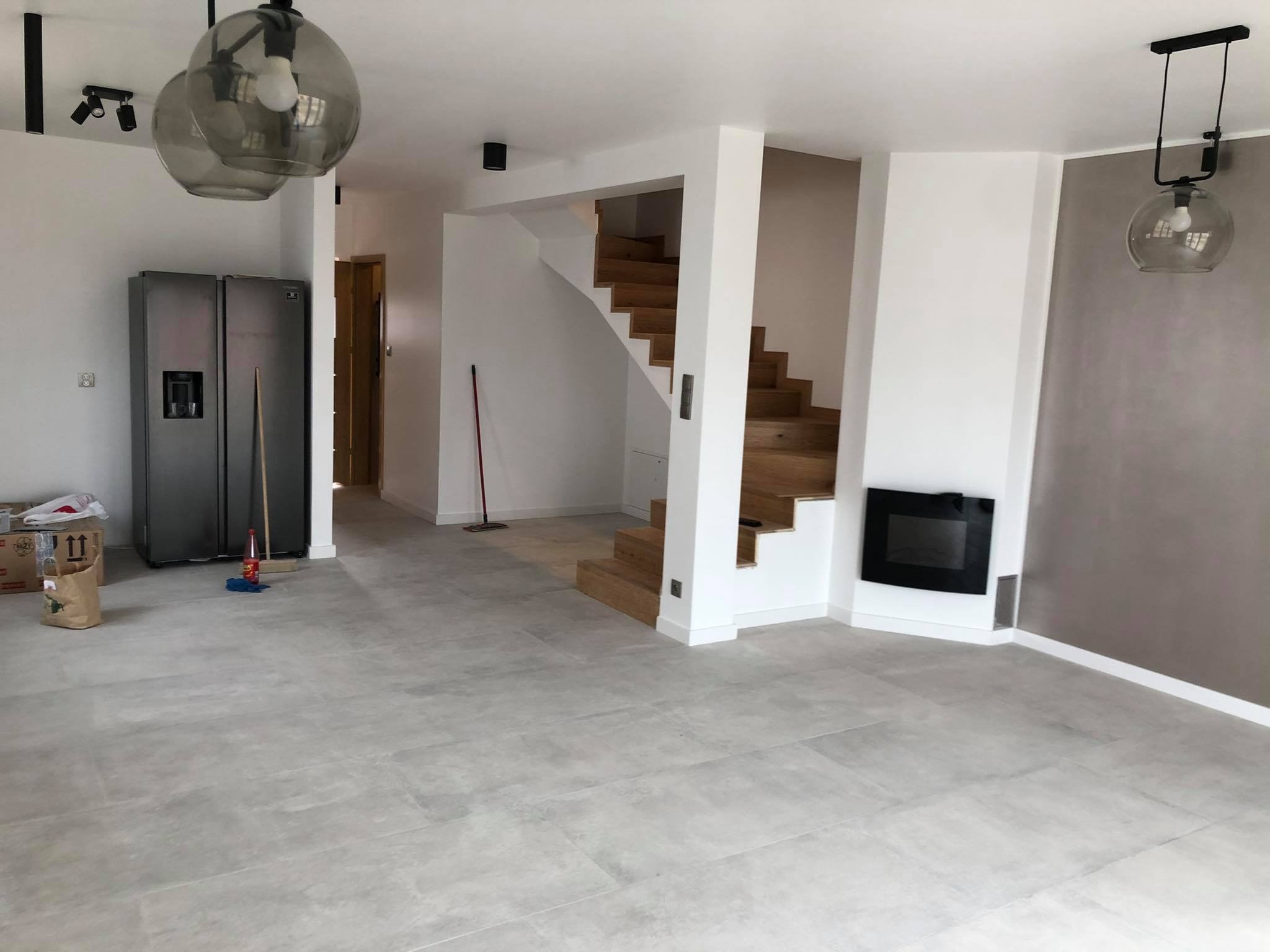Wnętrze salonu w trakcie wykańczania: szare płytki podłogowe, drewniane schody, białe ściany z fragmentem pomalowanym na szaro, nowoczesny kominek i szklane lampy wiszące.