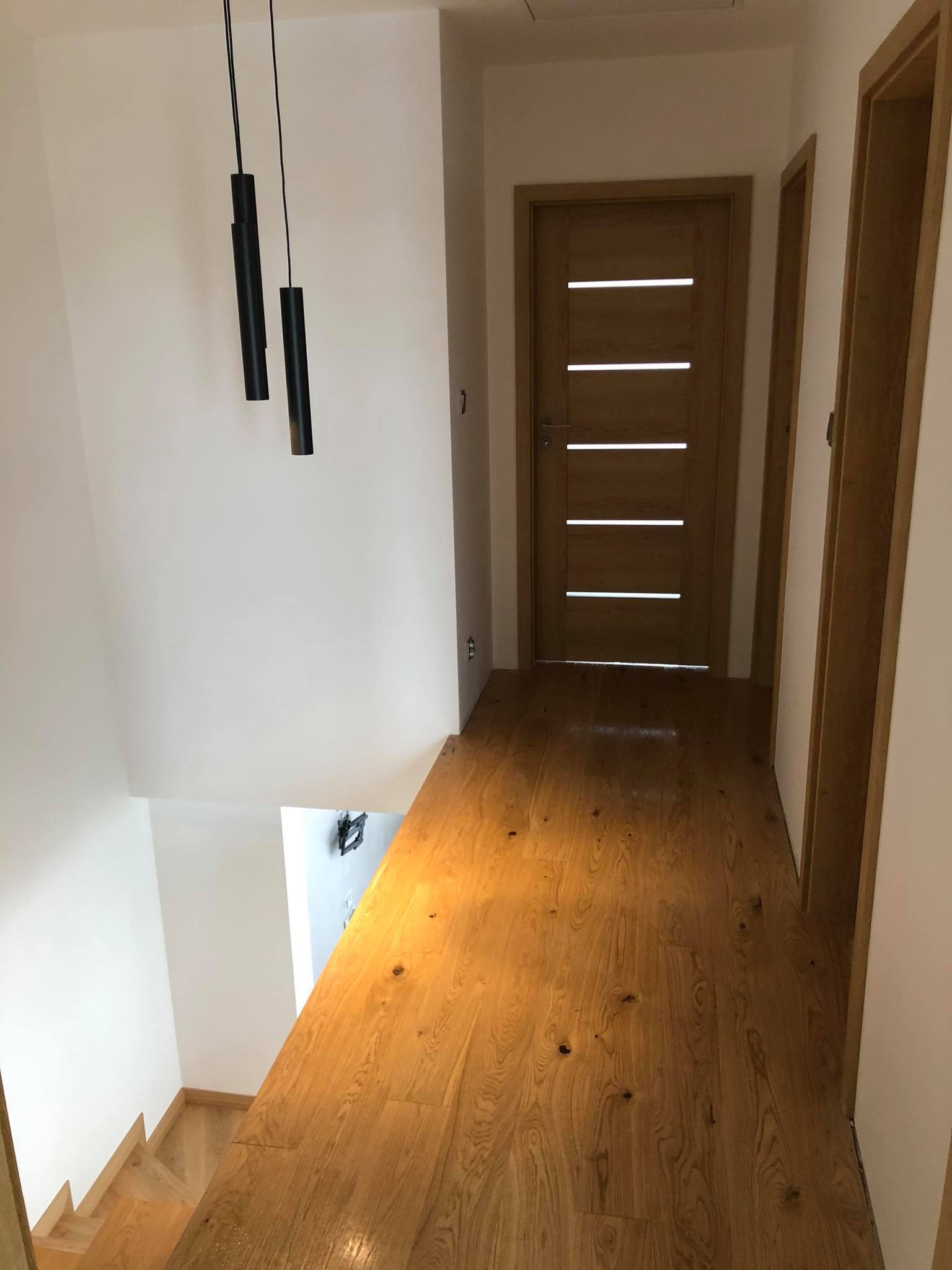 Korytarz z drewnianą podłogą, widoczne schody, drzwi z poziomymi przeszkleniami i wiszące czarne lampy.