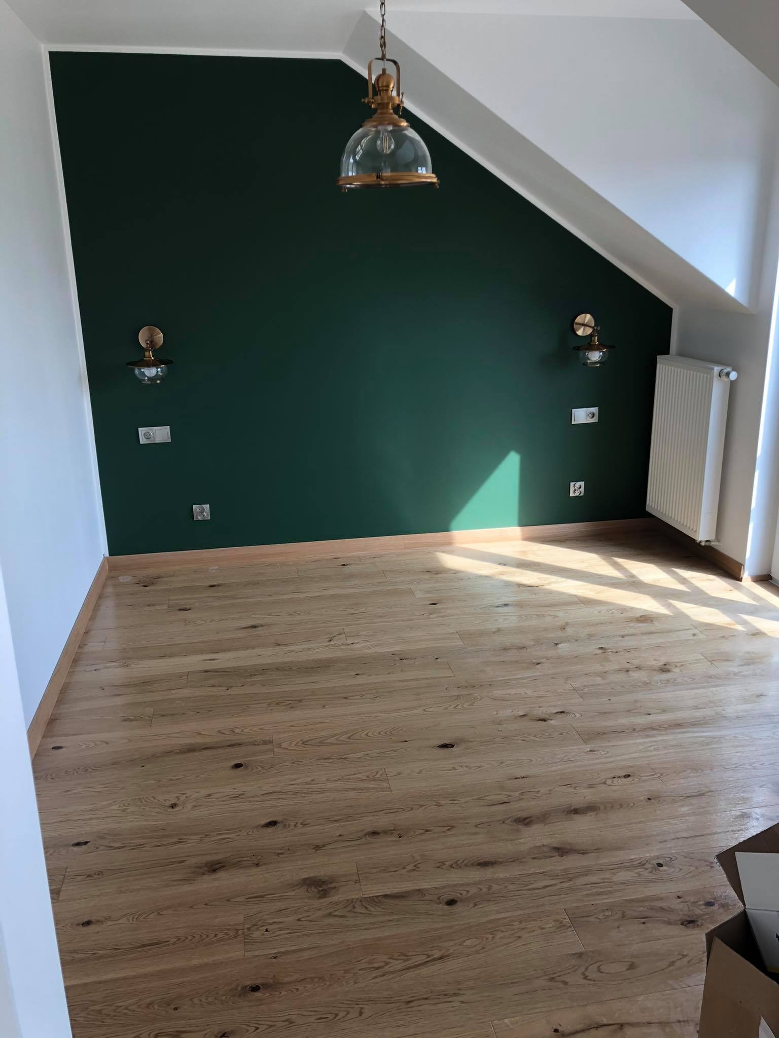 Pokój na poddaszu po remoncie: ciemnozielona ściana, drewniana podłoga, ozdobne lampy ścienne i wisząca, białe ściany boczne, grzejnik.