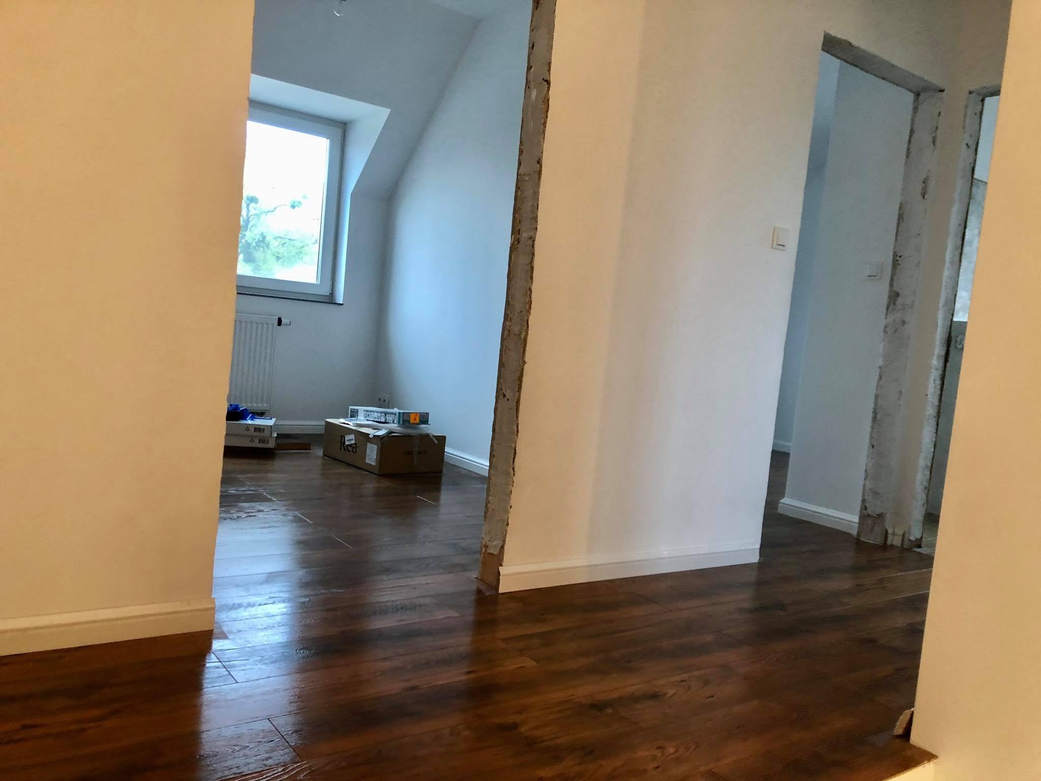 Wnętrze w trakcie remontu: brązowa podłoga z paneli, białe ściany, częściowo usunięta ścianka działowa z widocznym fragmentem konstrukcji, w tle okno w poddaszu i kartony z materiałami budowlanymi.