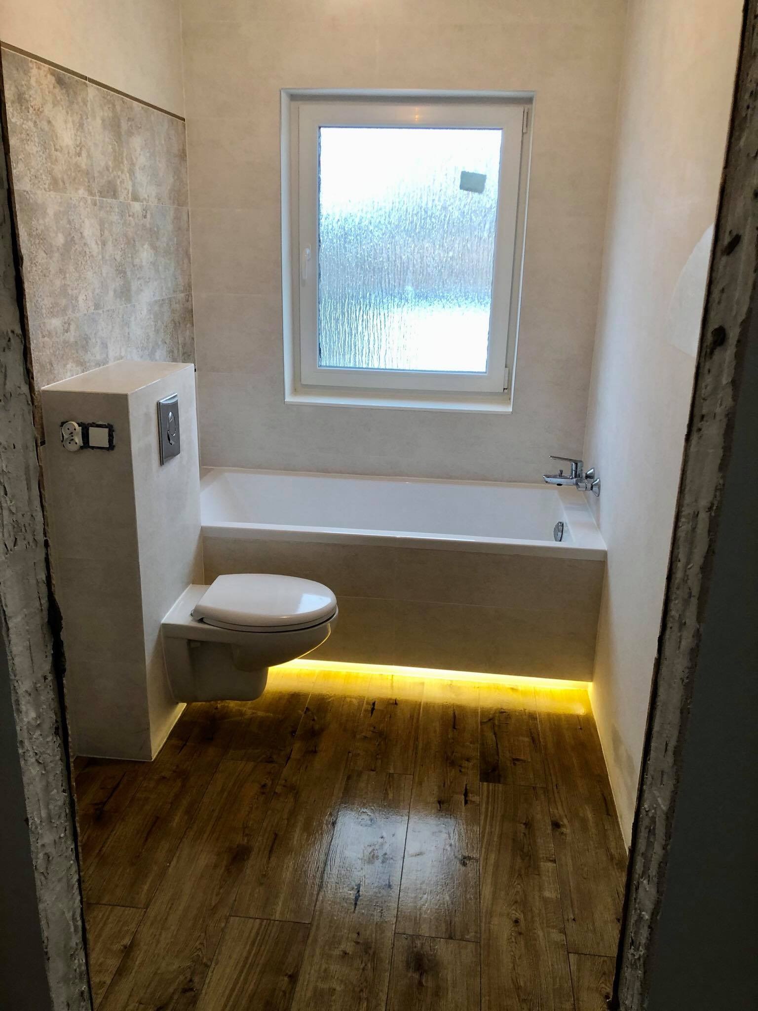 Wykończona łazienka z podświetloną wanną, drewnopodobną podłogą, podwieszaną toaletą i oknem z matową szybą.