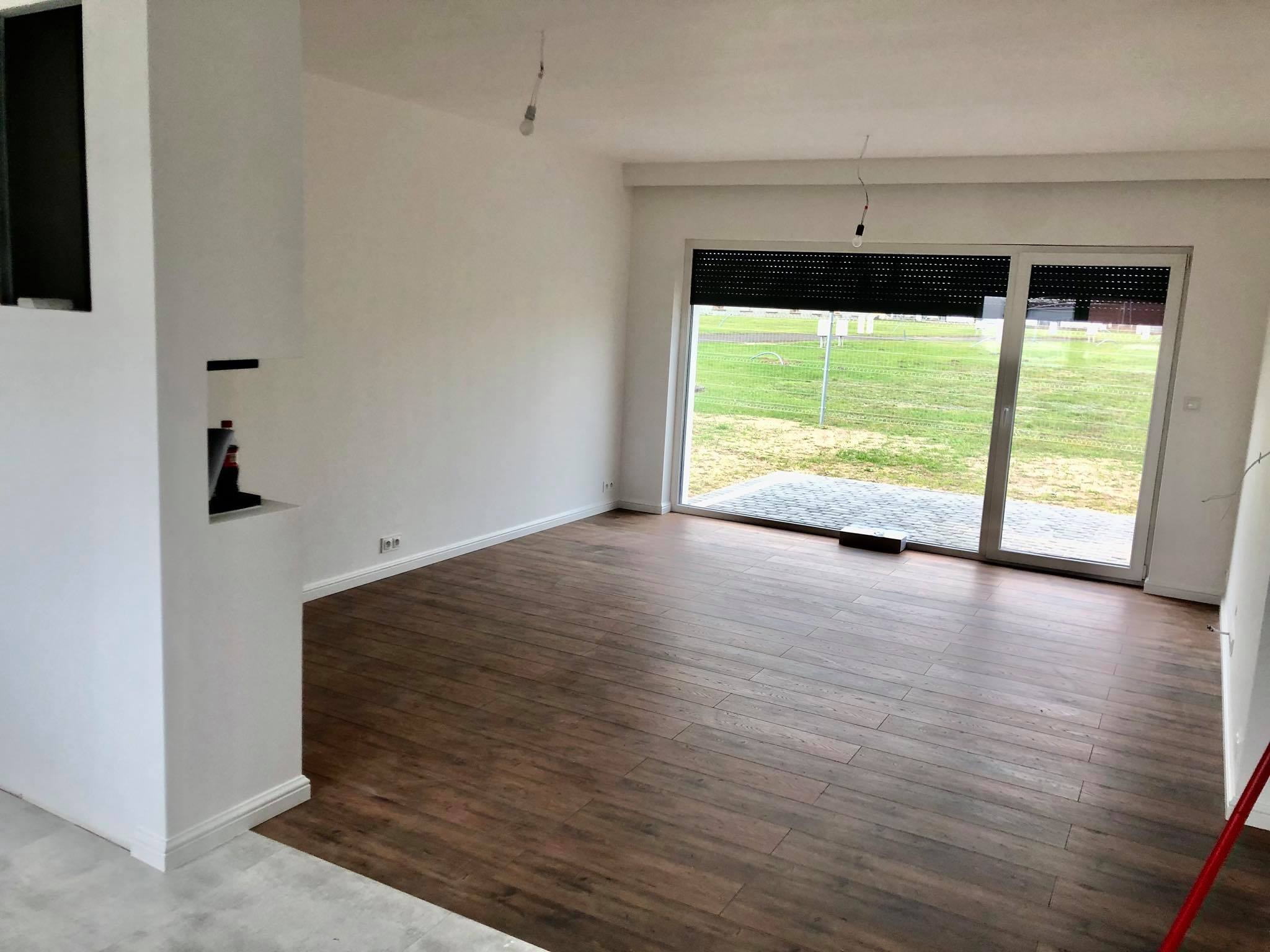 Wnętrze w trakcie wykańczania: brązowe panele podłogowe, białe ściany z widocznymi gniazdkami elektrycznymi, surowe oświetlenie i częściowo zamontowane rolety zewnętrzne.