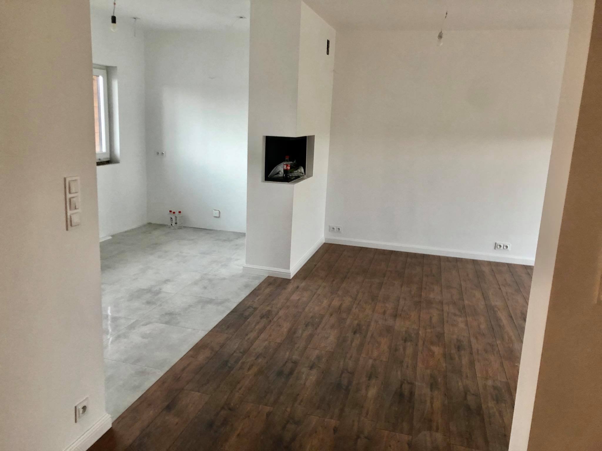 Wnętrze w trakcie wykańczania: połączenie ciemnej podłogi drewnopodobnej i jasnych, szarych płytek; białe ściany i kominek w surowym stanie.