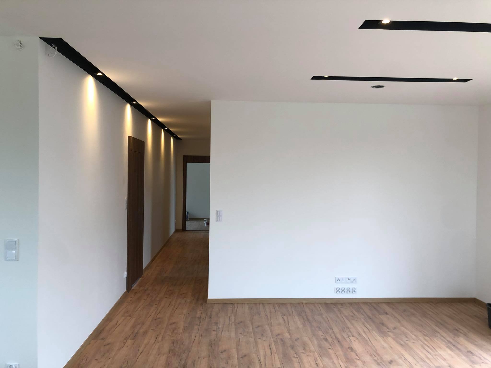 Jasne wnętrze z panelami podłogowymi, białymi ścianami i nowoczesnym oświetleniem sufitowym w postaci czarnych listew z punktowymi lampami LED.