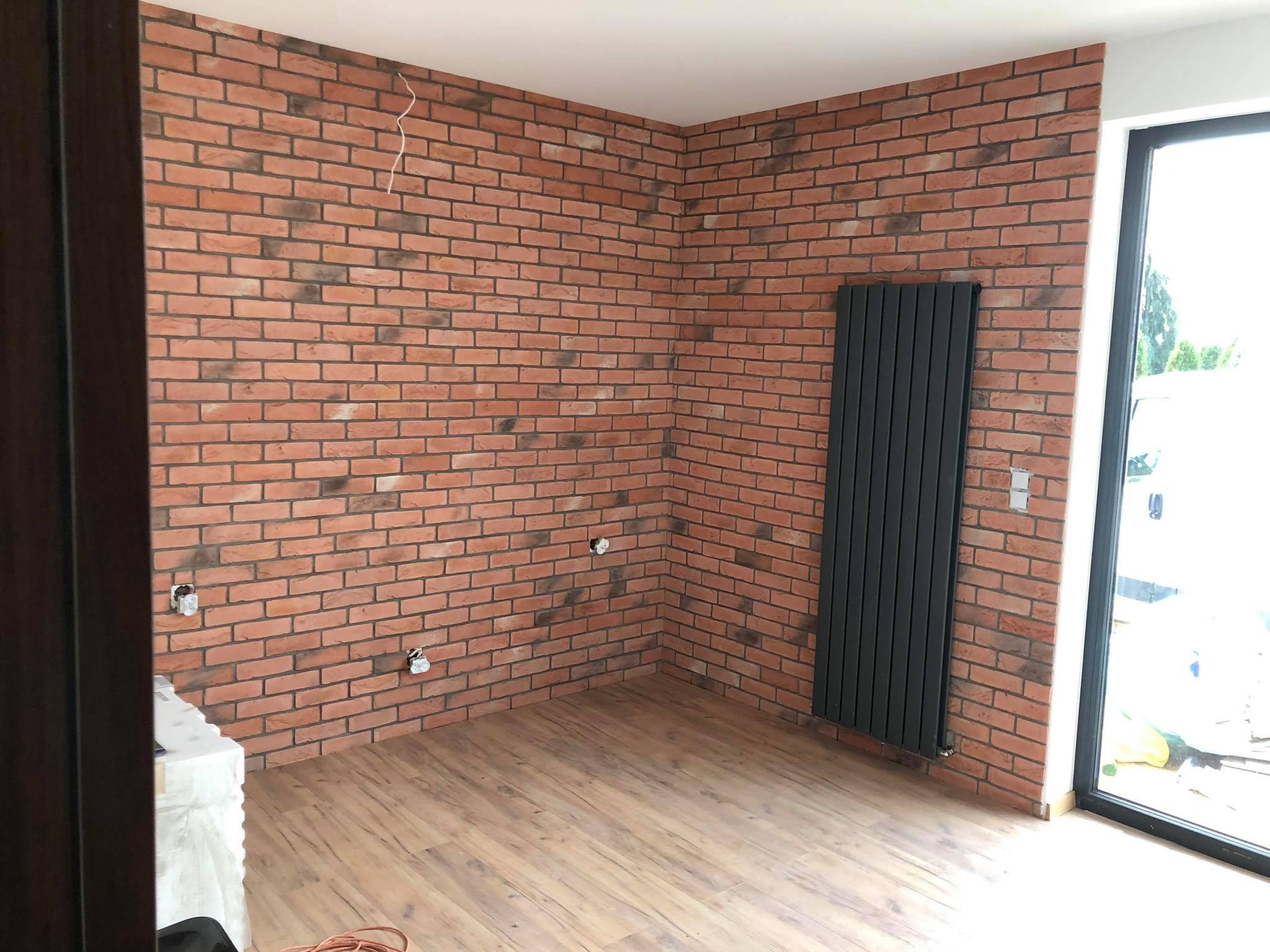 Ściana wykończona cegłą dekoracyjną, z zamontowanym grzejnikiem panelowym i widocznymi punktami elektrycznymi, podłoga wyłożona panelami imitującymi drewno, duże okno w czarnej ramie.