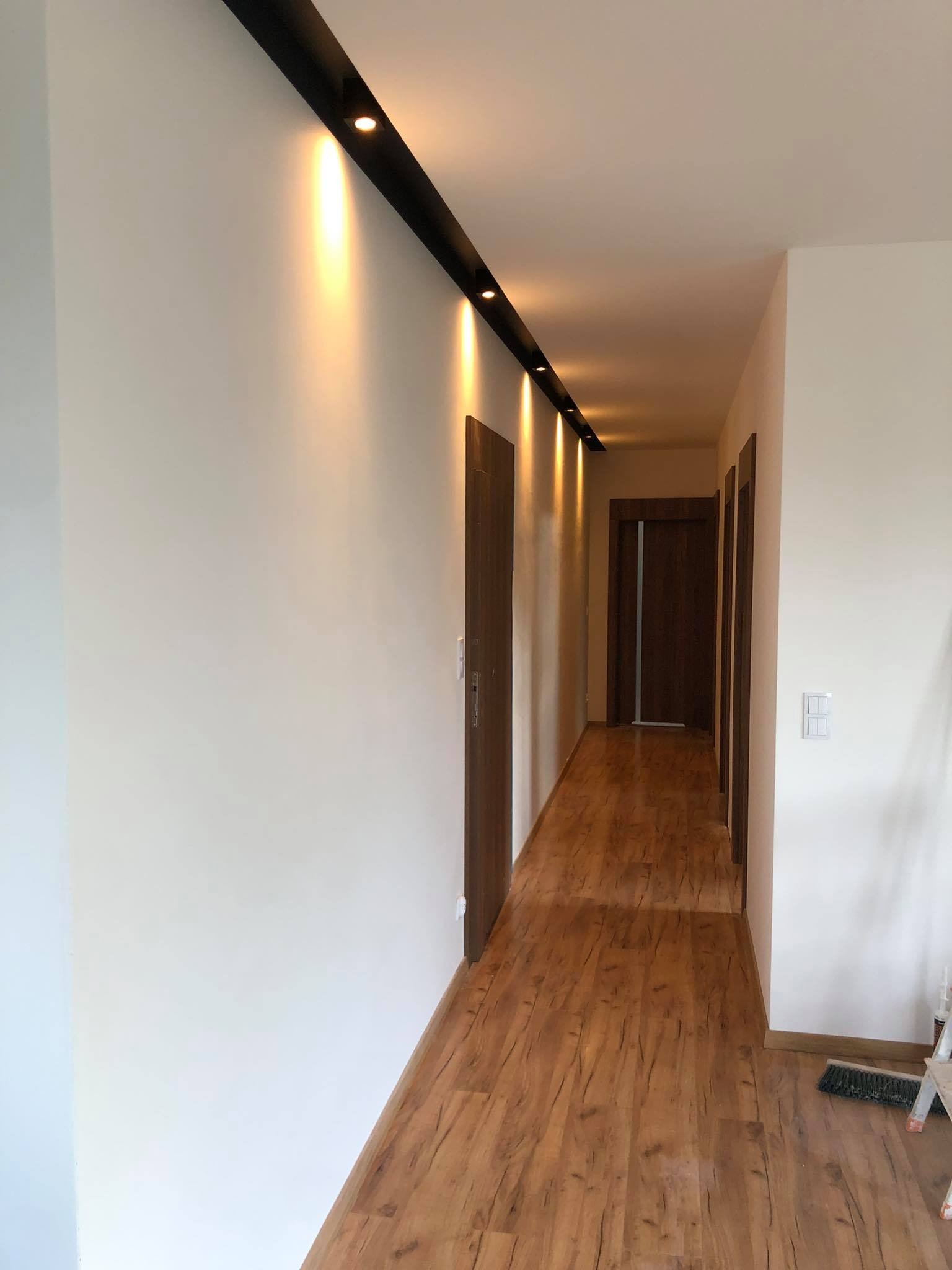 Korytarz z panelami podłogowymi imitującymi drewno, oświetlenie punktowe w czarnej listwie sufitowej, białe ściany, widoczne drzwi i włącznik światła, drabina oparta o ścianę.