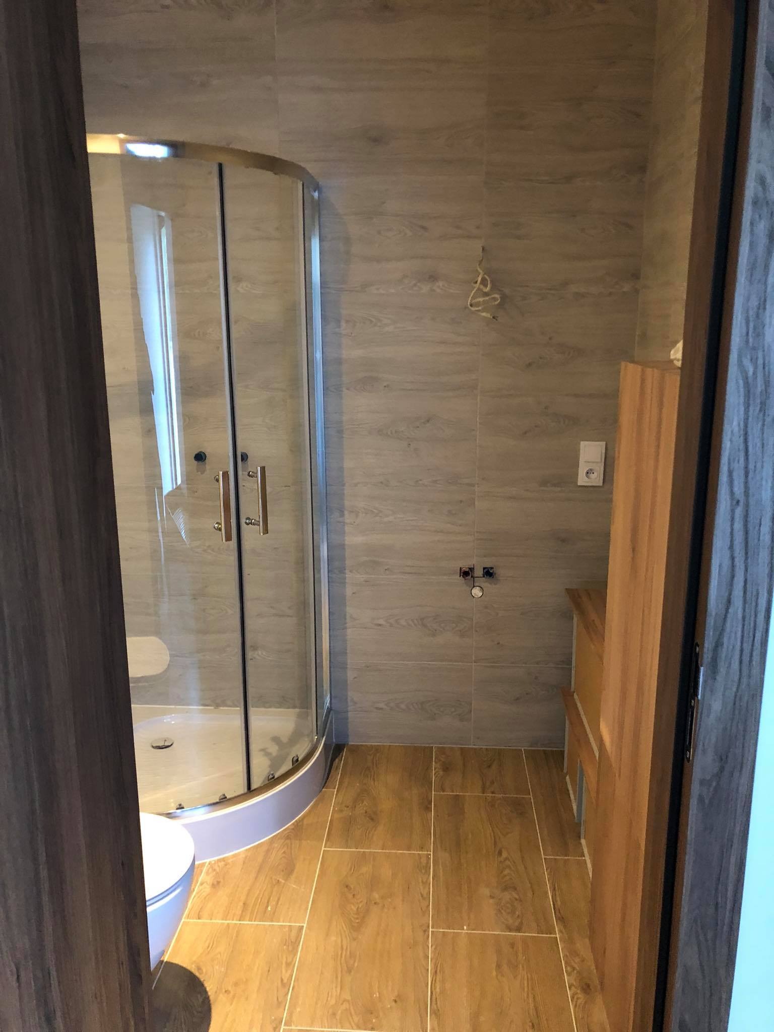 Wykończona łazienka z kabiną prysznicową z zaokrąglonymi drzwiami, płytkami imitującymi drewno na podłodze i ścianach oraz częściowo widoczną toaletą.