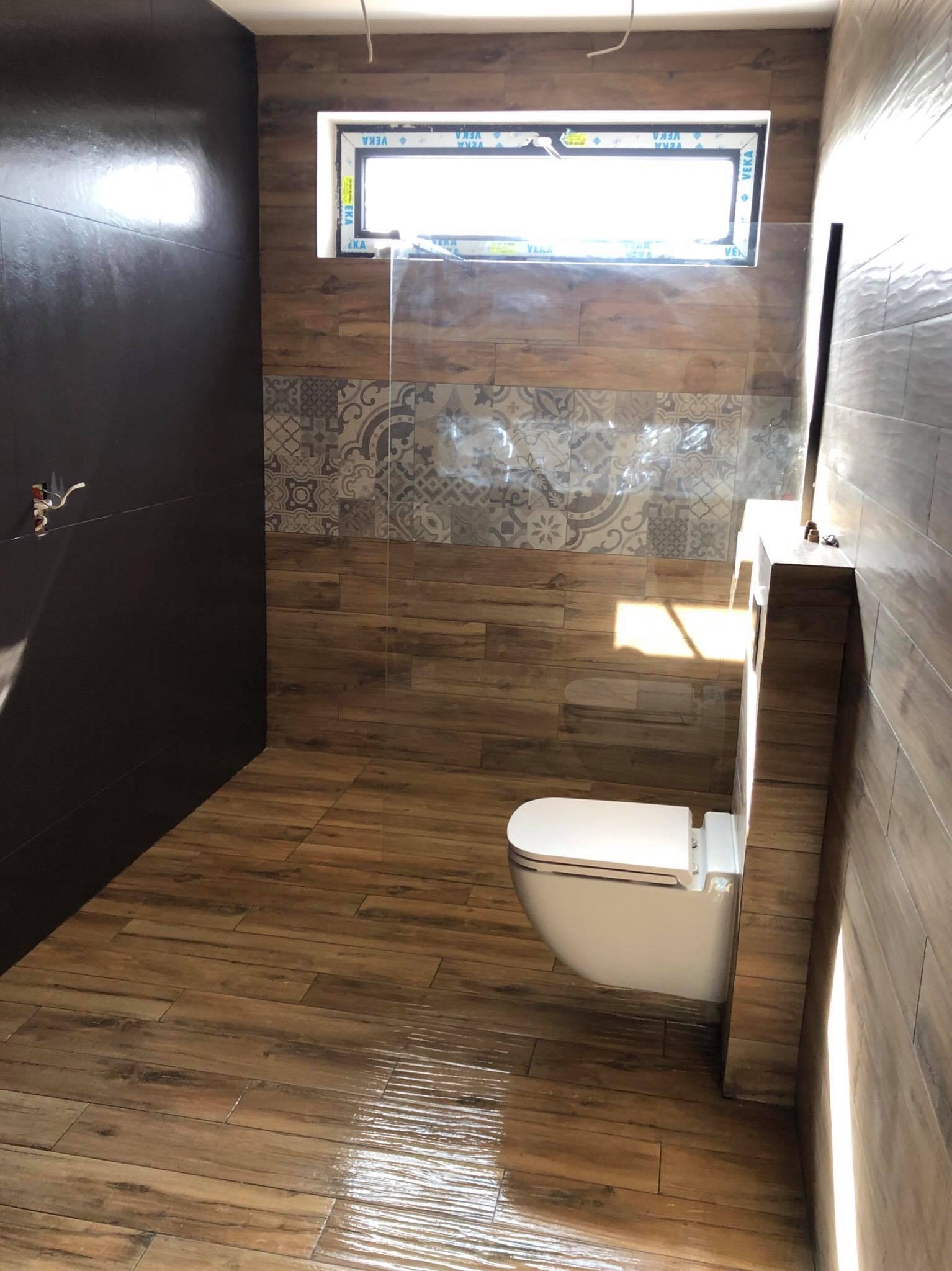 Nowoczesna łazienka z brązowymi płytkami imitującymi drewno, wisząca toaleta z zabudowaną spłuczką, okno z ramą PCV i częściowo przeszklona kabina prysznicowa.