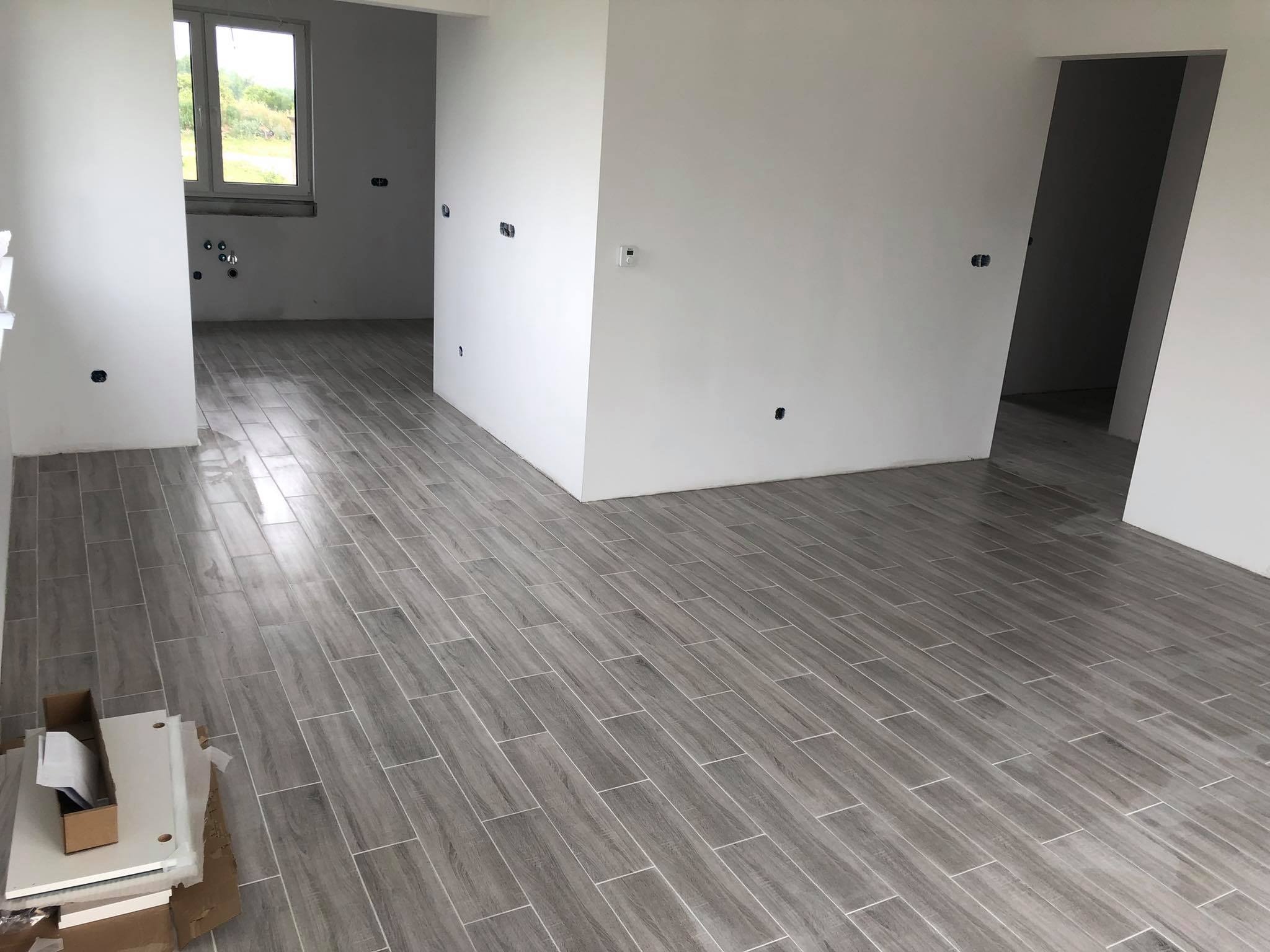 Szare płytki podłogowe imitujące drewno ułożone w jodełkę w surowym wnętrzu, widoczne przygotowane elementy meblowe w kartonach.