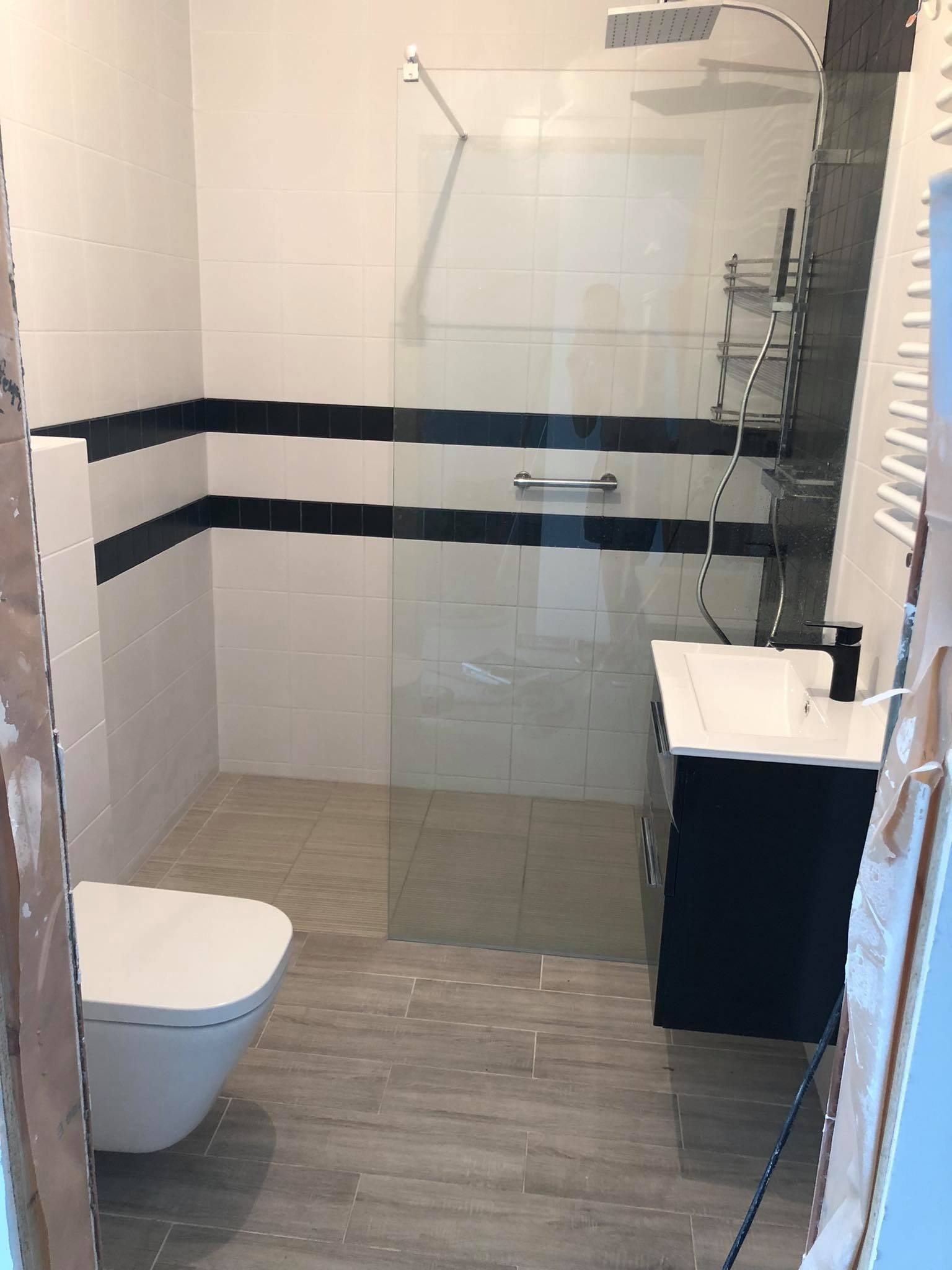 Nowoczesna łazienka z białymi i czarnymi płytkami, szklanym panelem prysznicowym, podwieszaną toaletą i ciemną szafką pod umywalką z czarną baterią, wykończona w szaro-beżowych płytkach podłogowych...