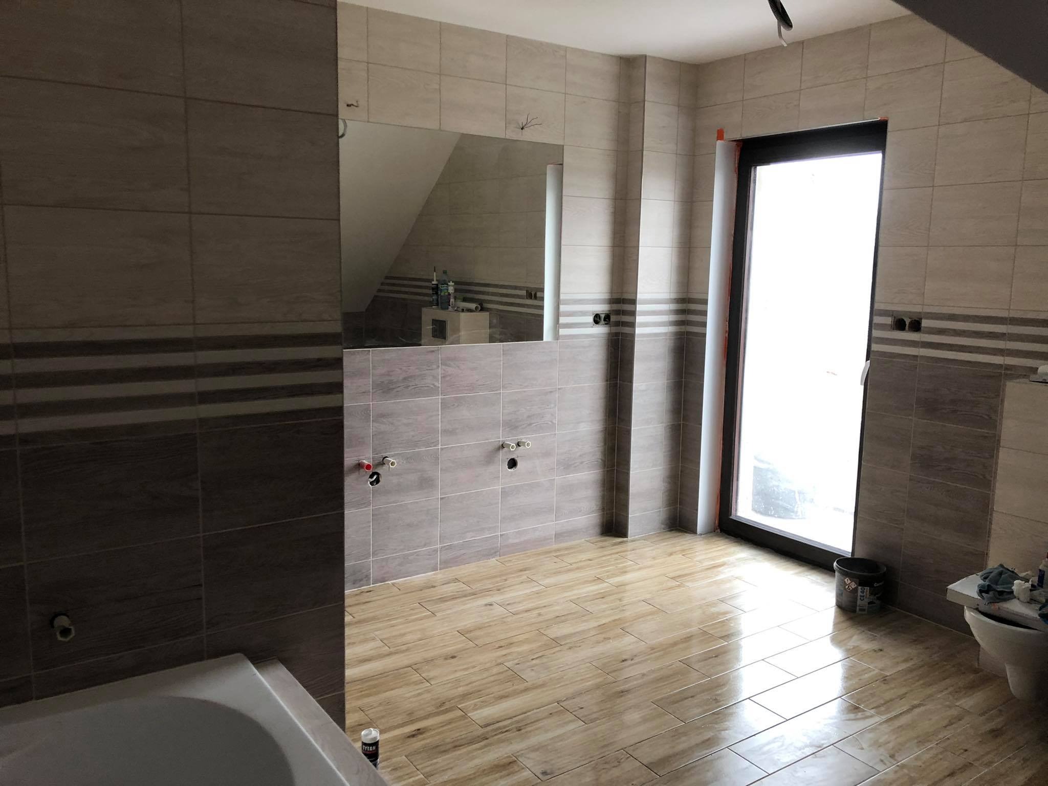 Wnętrze łazienki w trakcie remontu, widoczne położone płytki podłogowe imitujące drewno, częściowo wykończone ściany z szarych płytek z dekoracyjnym pasem, przygotowane podejścia pod umywalki...