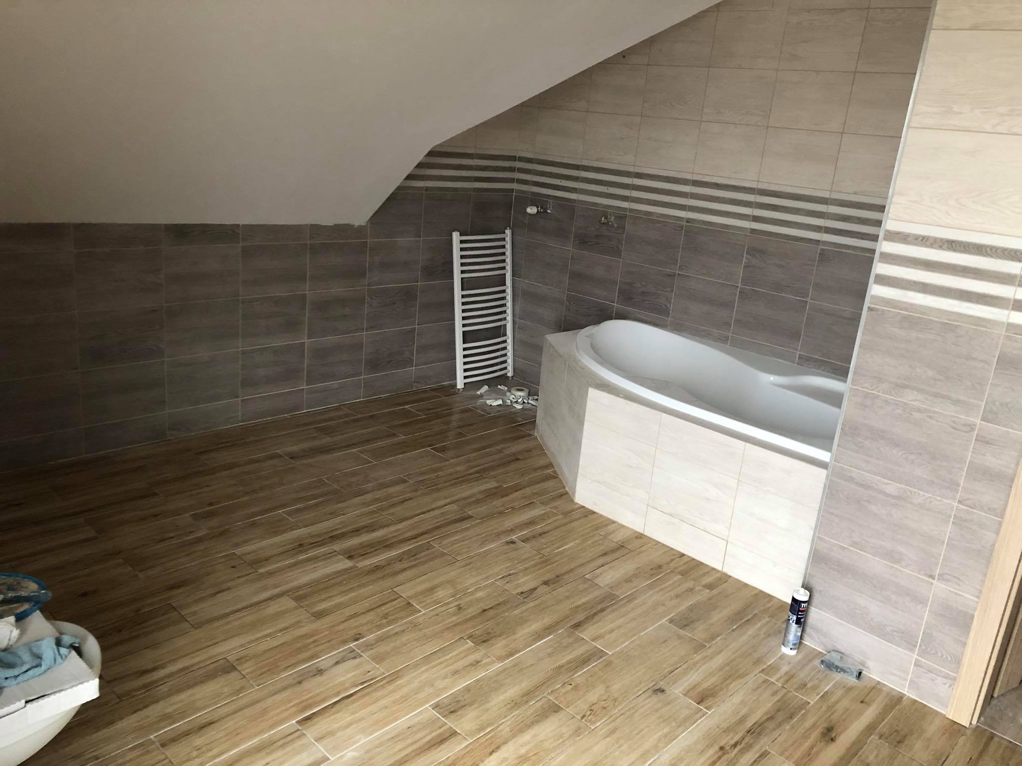 Łazienka na poddaszu z ułożonymi płytkami imitującymi drewno na podłodze, częściowo wykończona ściana z szarych płytek i biały grzejnik drabinkowy, wanna obudowana jasnymi płytkami.