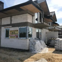 Surowy stan budynku jednorodzinnego w trakcie budowy, widoczne białe bloczki, okna z folią ochronną i reklama firmy budowlanej oferującej usługi budowlano-remontowe oraz tynki maszynowe, palety...