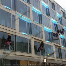 Pracownicy alpinistyczni myją okna na wysokościowym budynku biurowym z niebieskim oświetleniem LED, widoczne liny i sprzęt zabezpieczający.