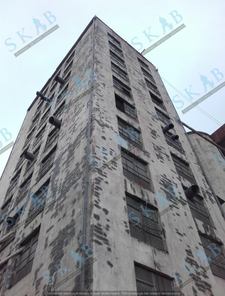 Wysoki, zaniedbany budynek przemysłowy z widocznymi śladami zabrudzeń i rurami wentylacyjnymi na elewacji, sfotografowany z dołu pod ostrym kątem.