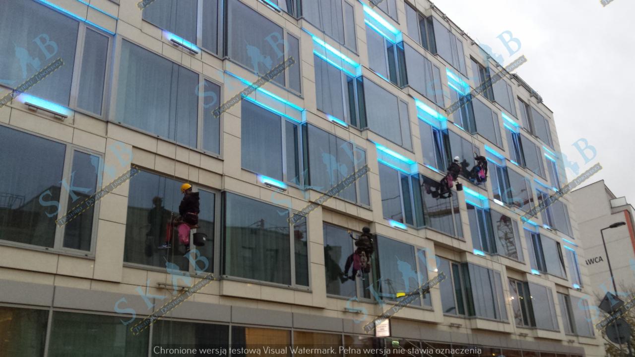 Pracownicy alpinistyczni myją okna na wysokościowym budynku biurowym z niebieskim oświetleniem LED, widoczne liny i sprzęt zabezpieczający.