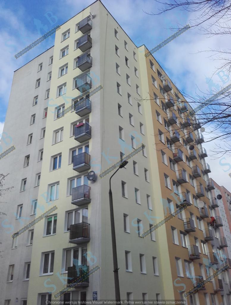 Wielopiętrowy blok mieszkalny z balkonami, częściowo ocieplony i pomalowany na różne odcienie beżu i szarości, widoczny z dołu pod kątem.