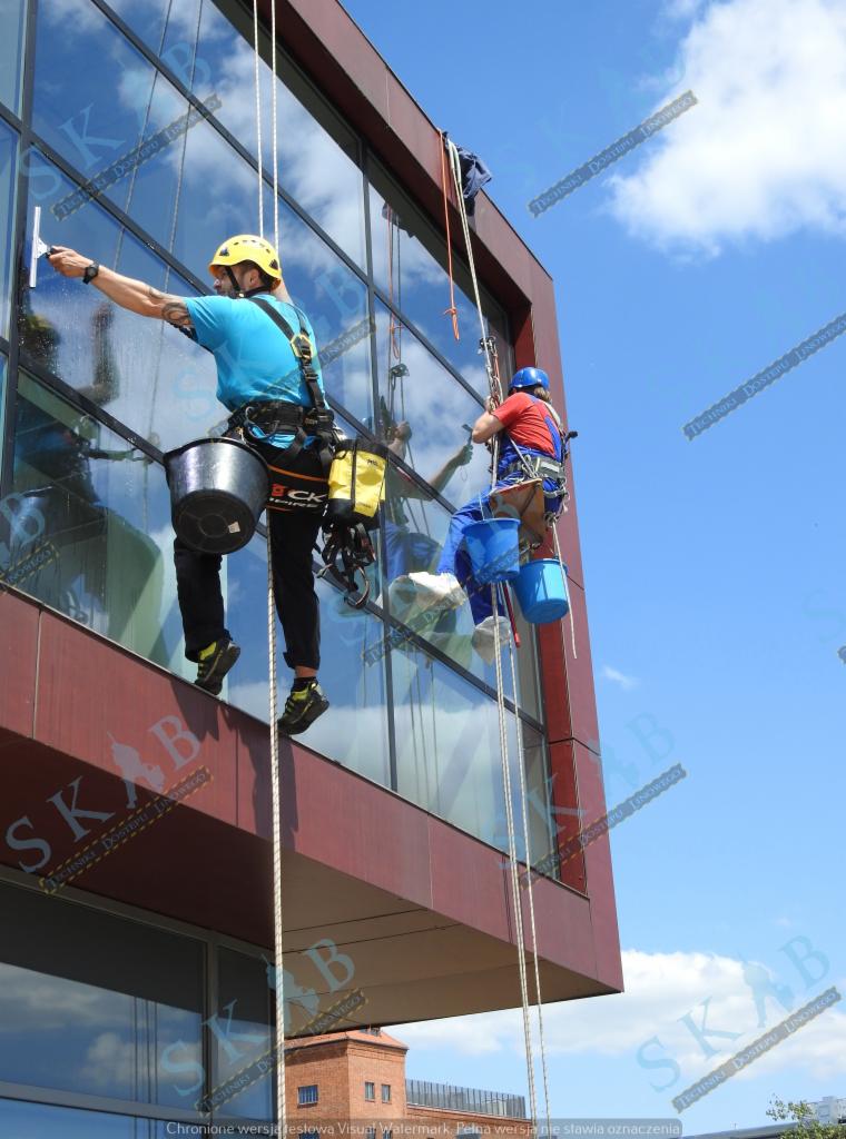 Dwóch alpinistów przemysłowych myje okna wieżowca, jeden z żółtym kaskiem używa ściągaczki, drugi w niebieskim kasku opuszcza się na linie, widoczne odbicie nieba w oknach.