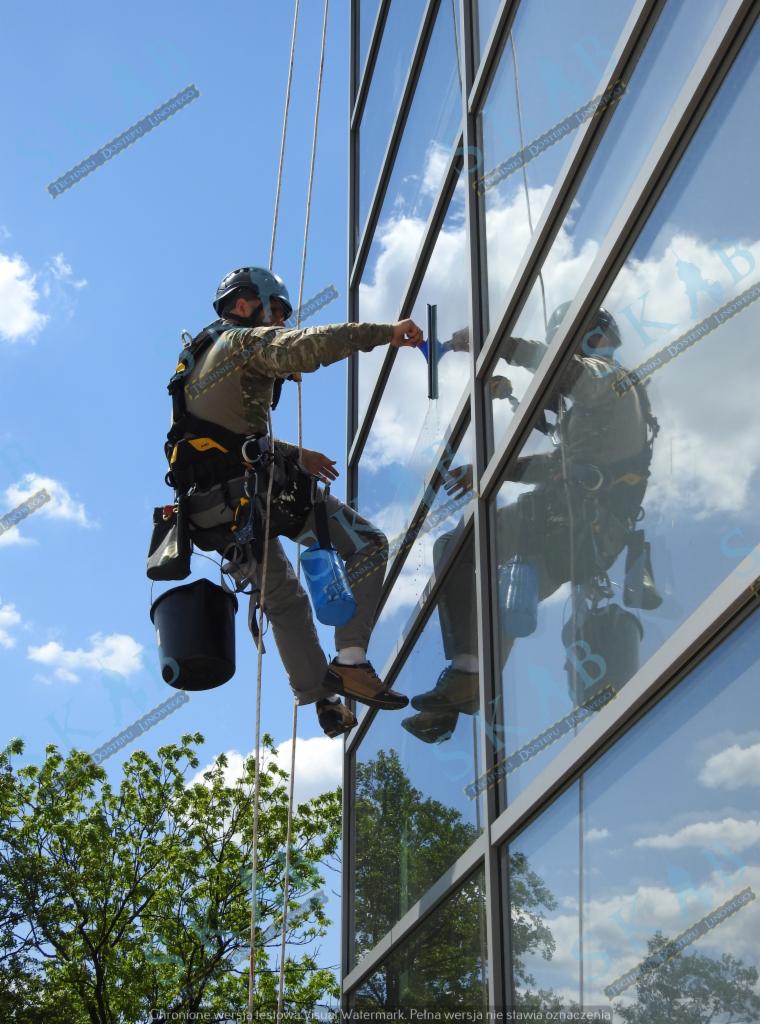 Pracownik w uprzęży, myjący okna wieżowca za pomocą ściągaczki, z widocznym odbiciem w szybach i błękitnym niebem w tle.